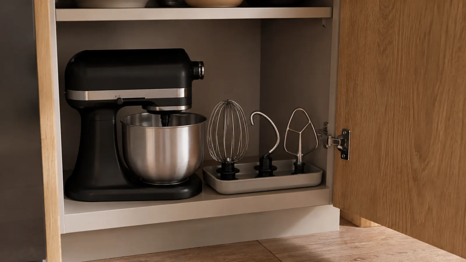 Small stand mixer stored on a shelf next to its whisk and beater attachments in a shallow cabinet