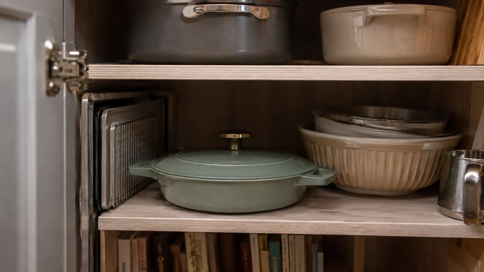 Enameled braiser stacked on a narrow cabinet shelf between a sheet pan and a stack of bowls