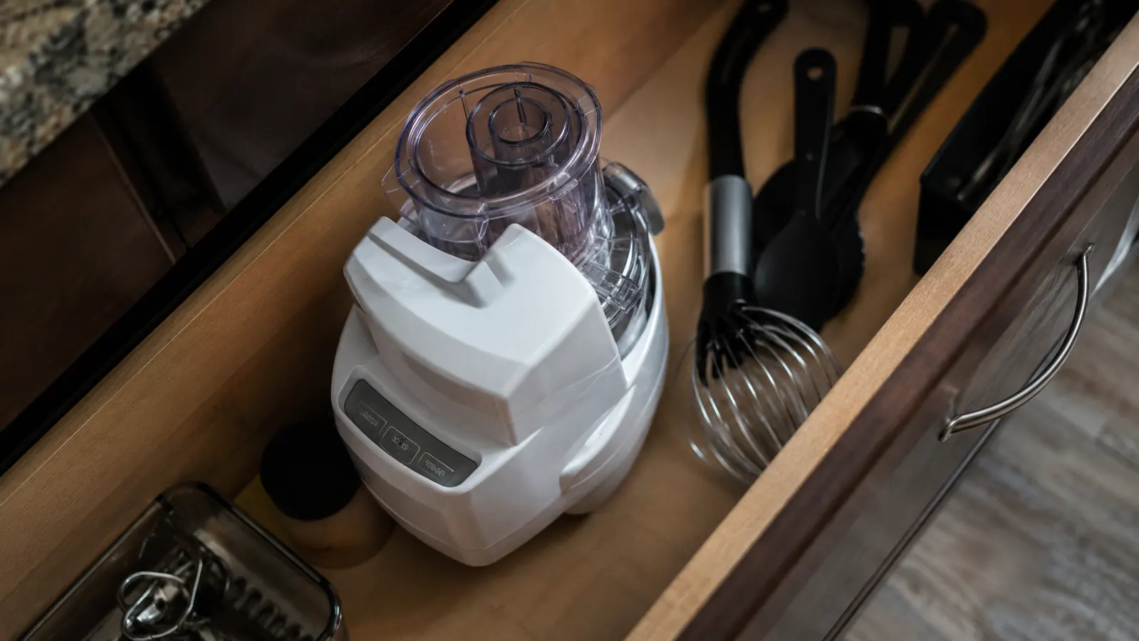 Small white food processor bowl and base standing upright in a narrow kitchen drawer next to cooking utensils