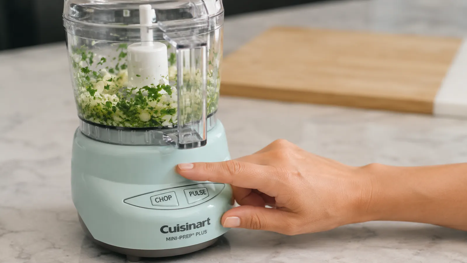 A hand pressing the pulse button on the Cuisinart Mini-Prep Plus with garlic and parsley chopped inside the bowl