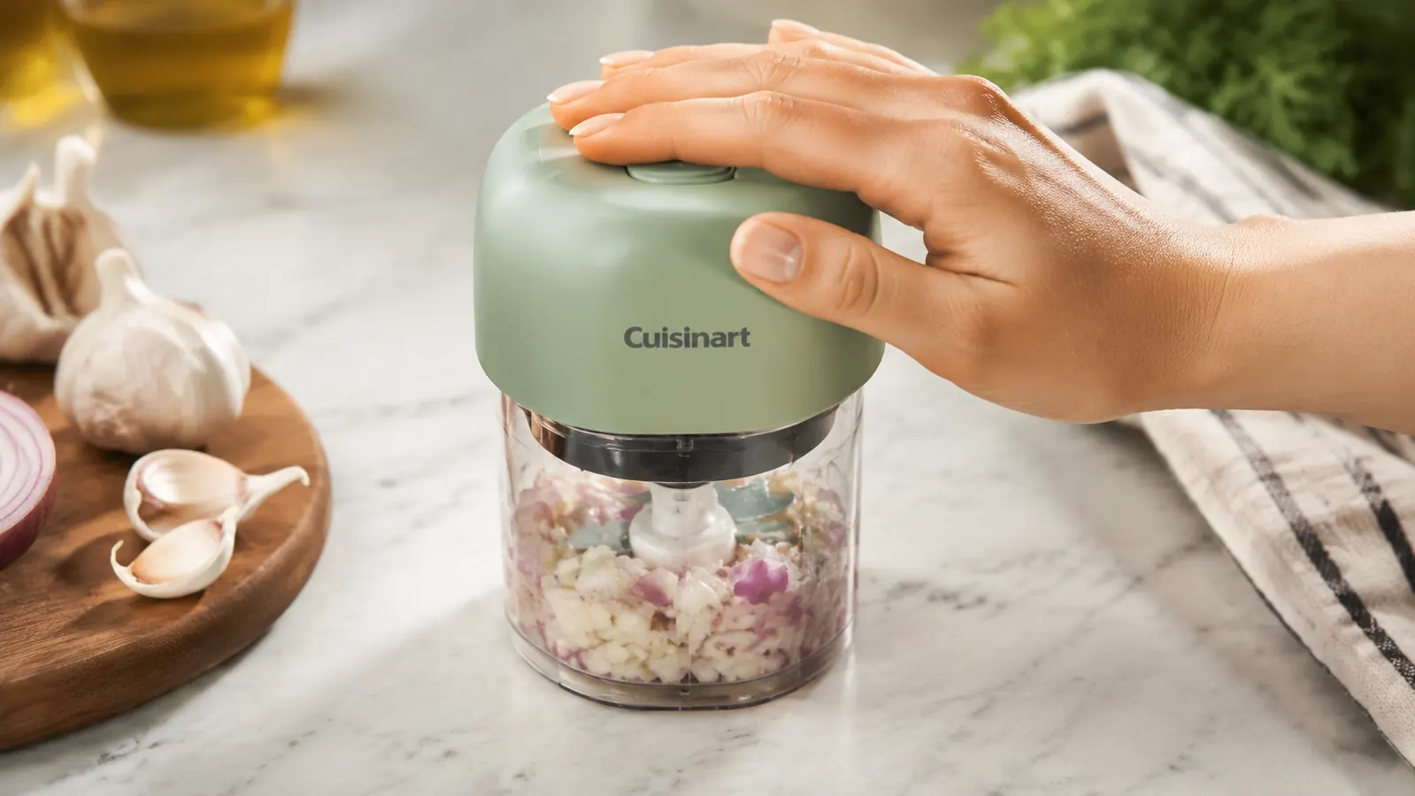 Hand pressing the pulse button on the Cuisinart Mini-Prep Plus with garlic cloves visible inside the clear plastic bowl