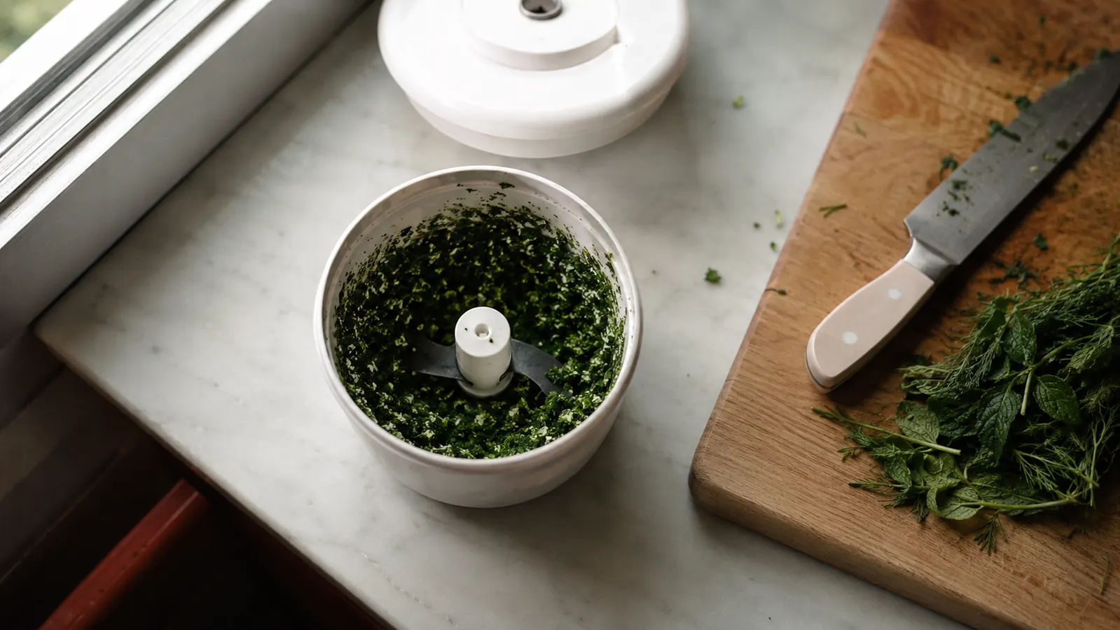 Overhead view of a small food processor bowl full of finely chopped herbs on a narrow kitchen counter