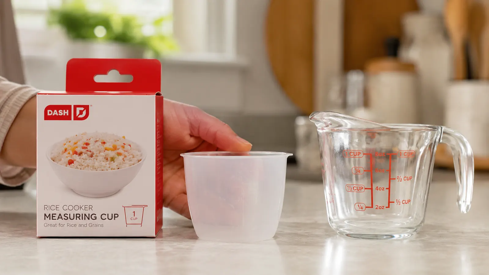 Hand holding the small plastic measuring cup that comes with the Dash Mini Rice Cooker next to a standard glass measuring cup, showing the size difference