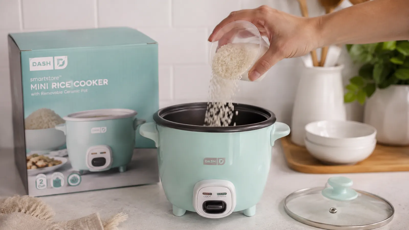 Hand pouring measured white rice from a cup into the removable ceramic pot of the Dash Mini Rice Cooker