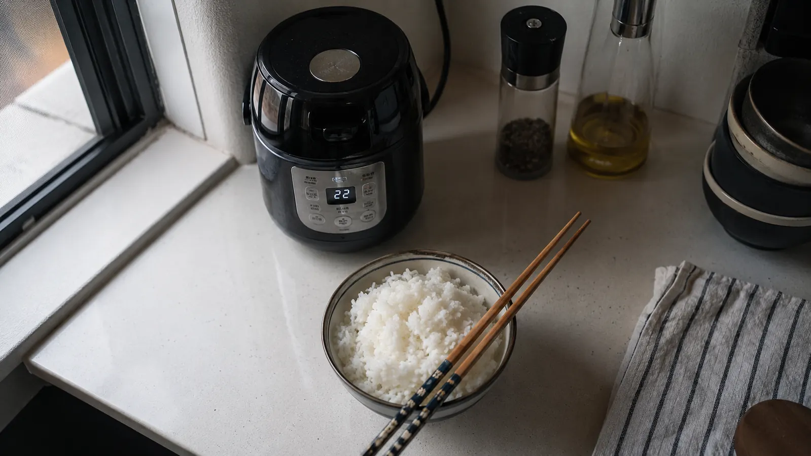 Overhead view of a small bowl of finished rice next to a compact rice cooker on a narrow kitchen counter