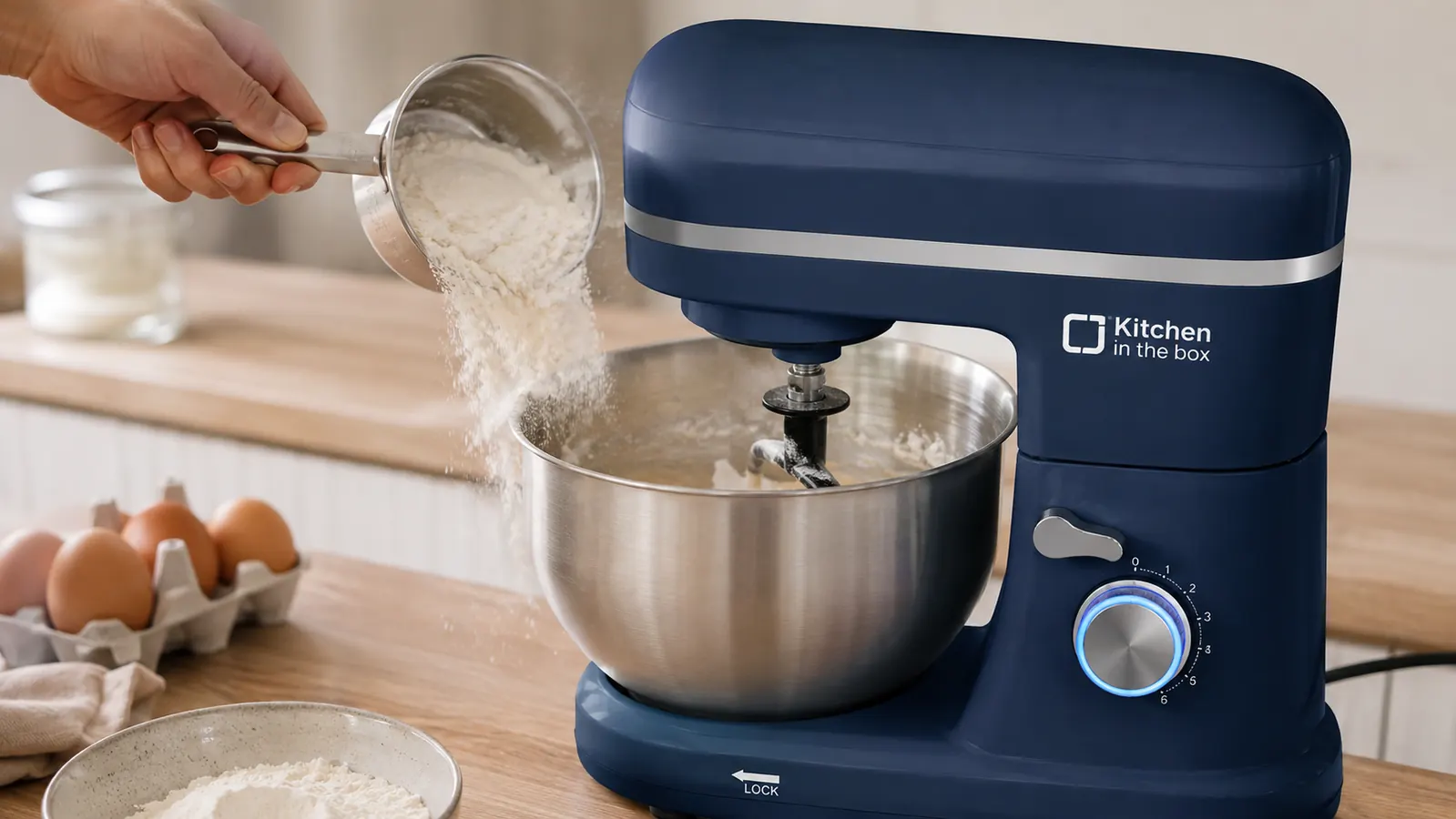 A hand adding flour to the mixing bowl of a small stand mixer while the flat beater runs on a low speed