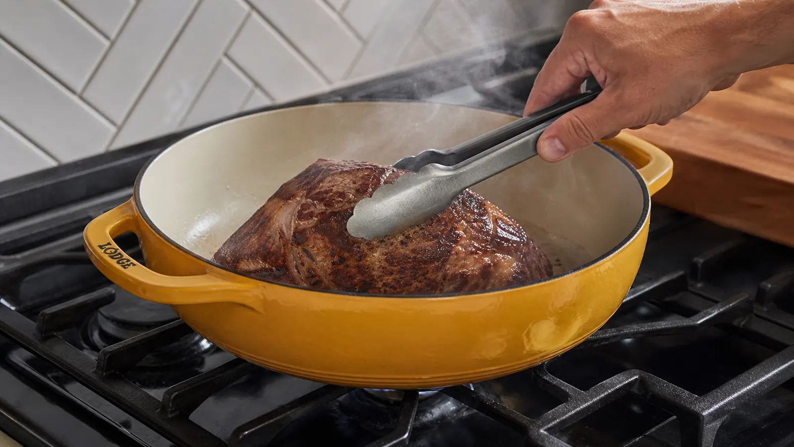 A hand searing a chuck roast in a wide enameled braiser on the stovetop