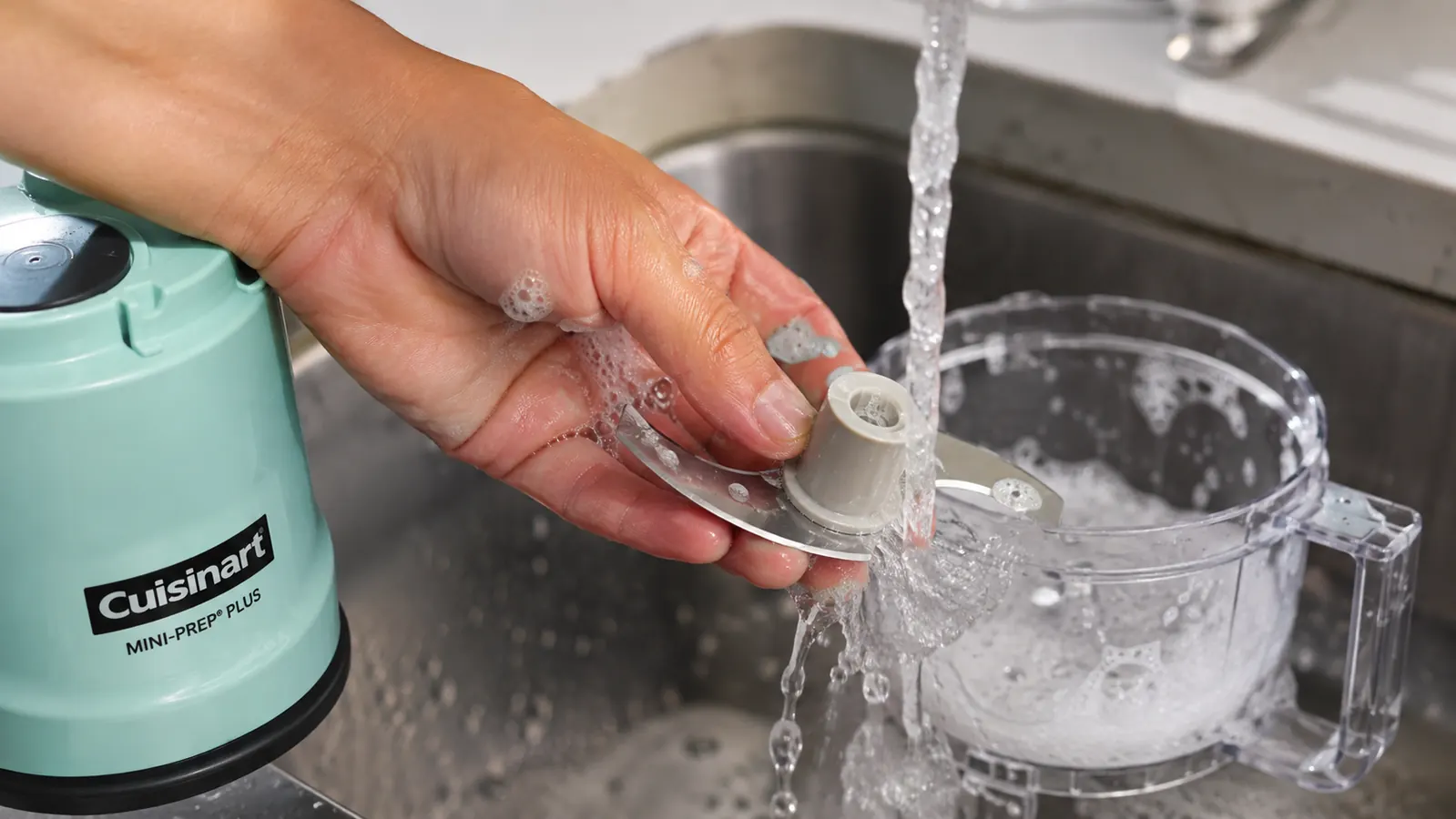 A stainless steel blade and small clear work bowl of a mini food processor being wiped clean at a kitchen sink