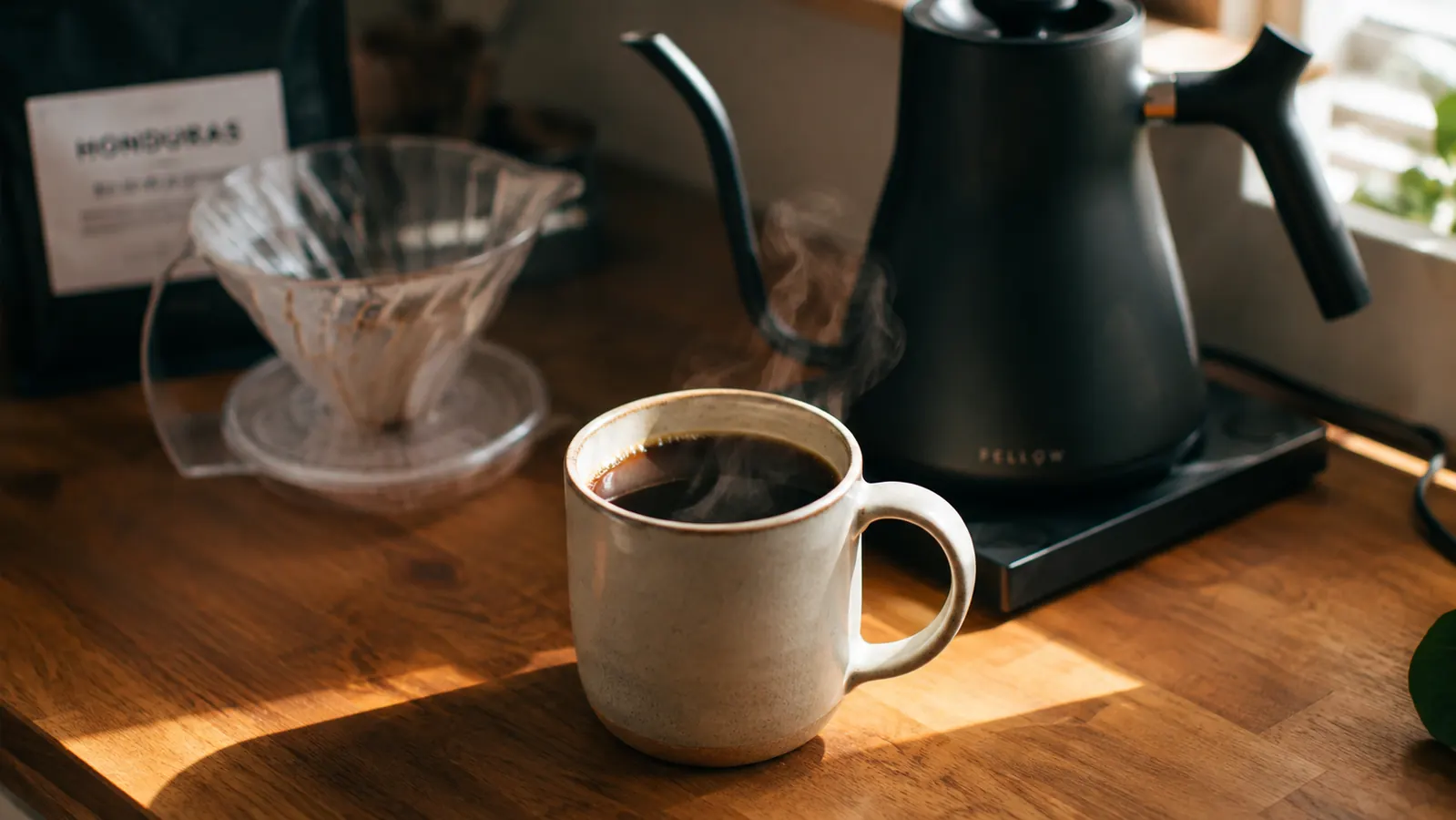 A finished mug of pour-over coffee sitting next to a gooseneck kettle on a small kitchen counter in morning light