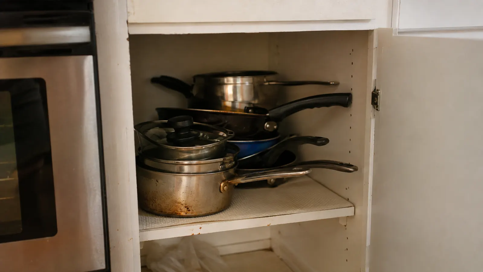 A small, crowded kitchen cabinet with mismatched pots and pans stacked on top of each other