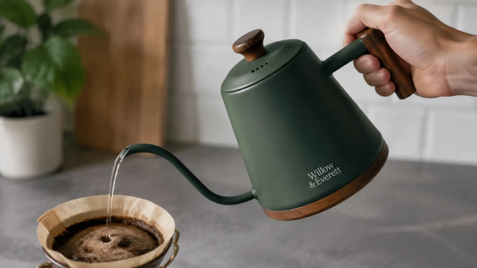 Hand holding the gooseneck kettle by its handle, pouring a thin controlled stream into a coffee filter